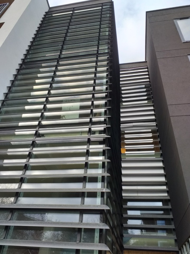 Wysoki budynek z fasadą z poziomych, metalowych żaluzji zamontowanych na oknach, widok z dołu w pochmurny dzień.