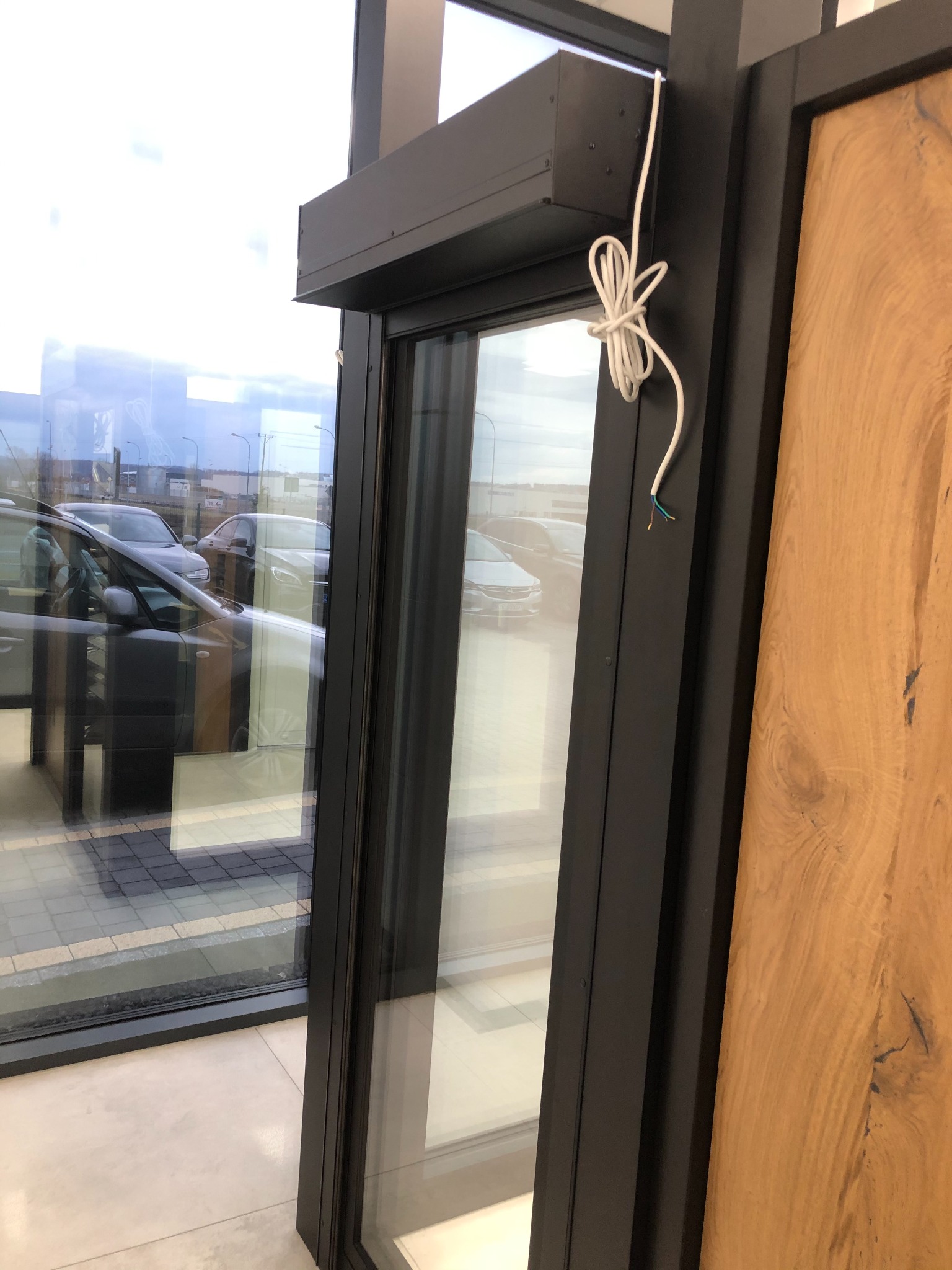 Ciemna rama okna PCV z roletą zewnętrzną w trakcie montażu, widoczne kable elektryczne, na zewnątrz zaparkowane samochody.