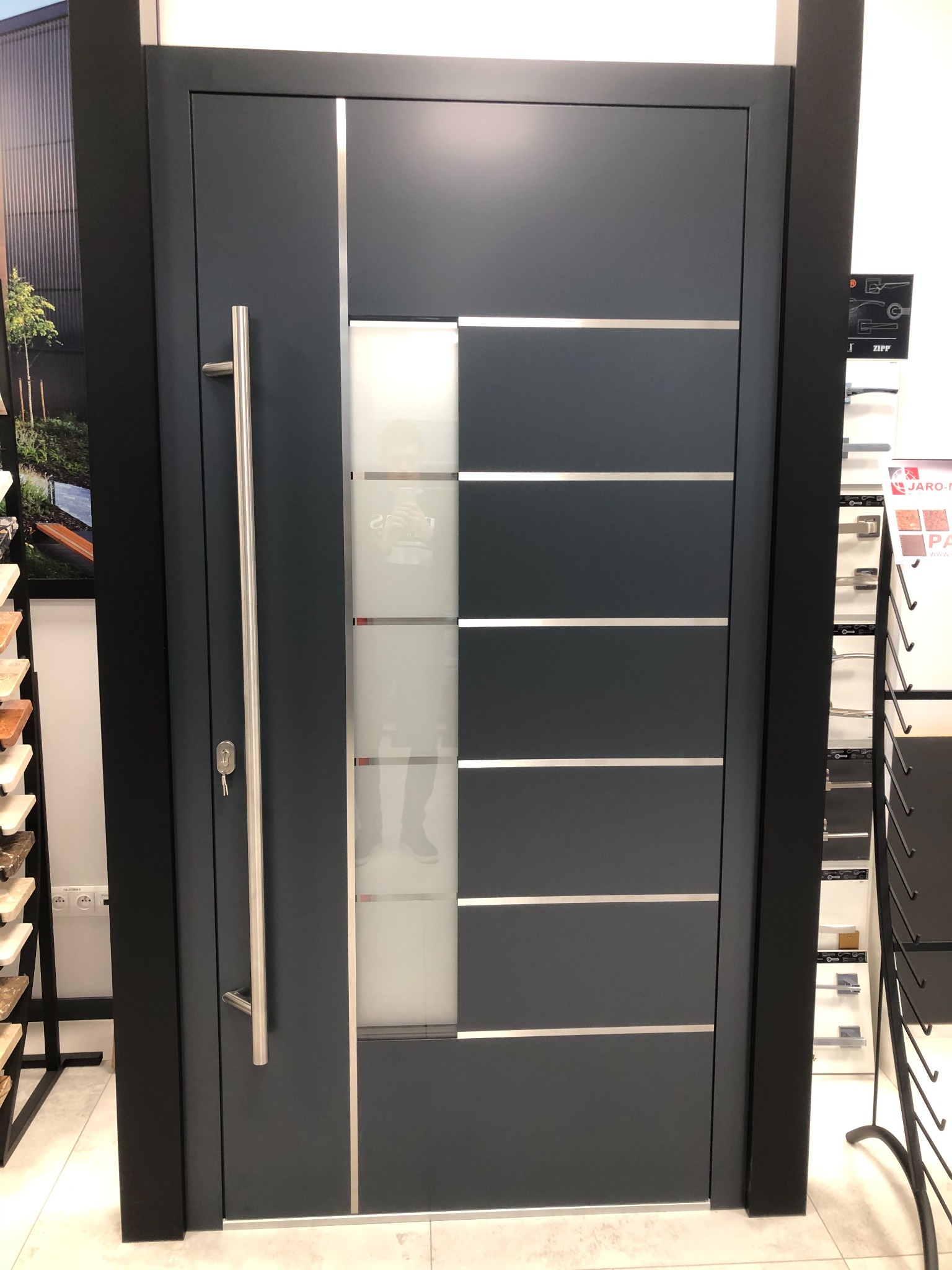 Nowoczesne, antracytowe drzwi wejściowe z długim, pionowym uchwytem i przeszklonym panelem z poziomymi, srebrnymi akcentami, prezentowane w salonie sprzedaży.