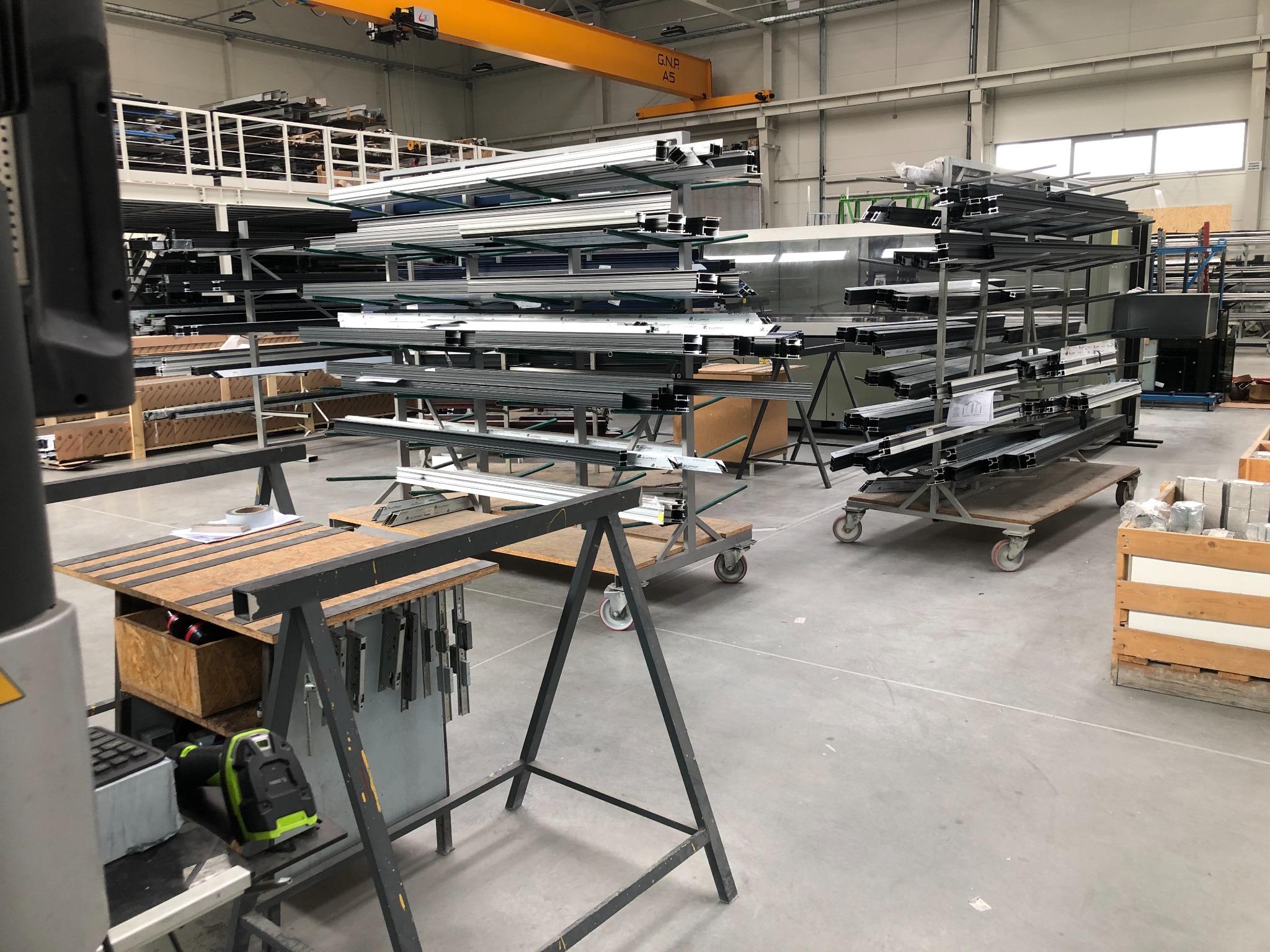 Magazyn profili aluminiowych do okien i parapetów, widoczne wózki transportowe, stanowisko pracy z narzędziami i materiałami.