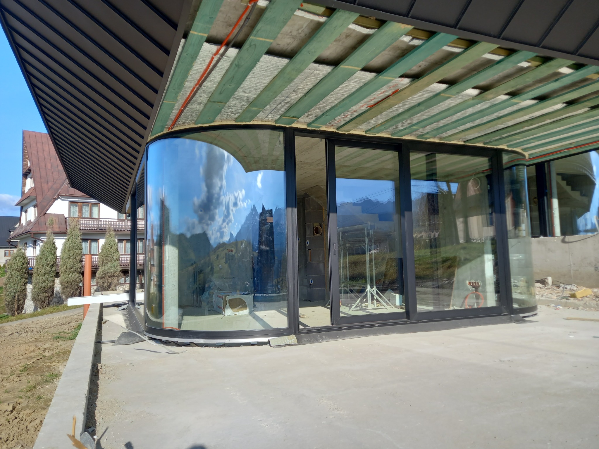 Panoramiczne, aluminiowe okna z zaokrąglonym narożnikiem w nowo budowanym domu, z widokiem na góry odbijającym się w szybach.