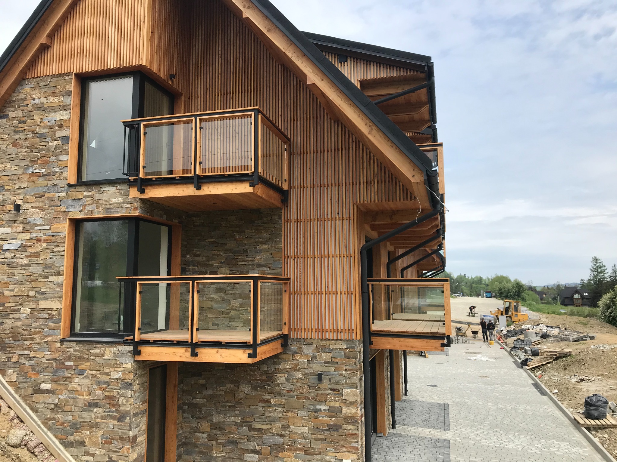 Nowoczesny budynek w górskim stylu z kamienną elewacją, drewnianymi elementami i balkonami z szklanymi balustradami, widoczne prace wykończeniowe na zewnątrz.