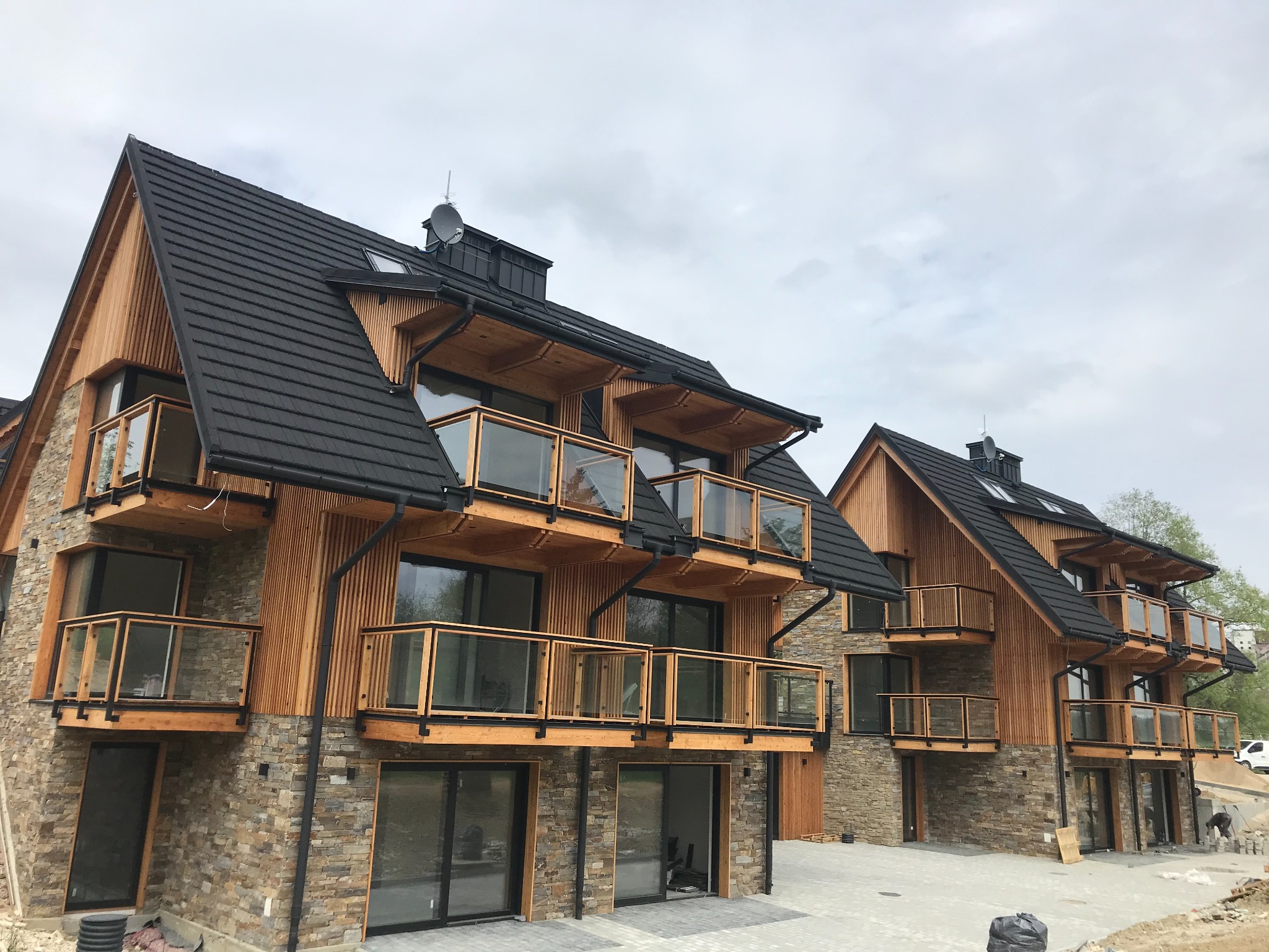 Nowoczesny budynek apartamentowy w stylu góralskim z drewnianą elewacją, kamiennym cokołem i balkonami z szklanymi balustradami, z czarną dachówką i widoczną anteną satelitarną na dachu.