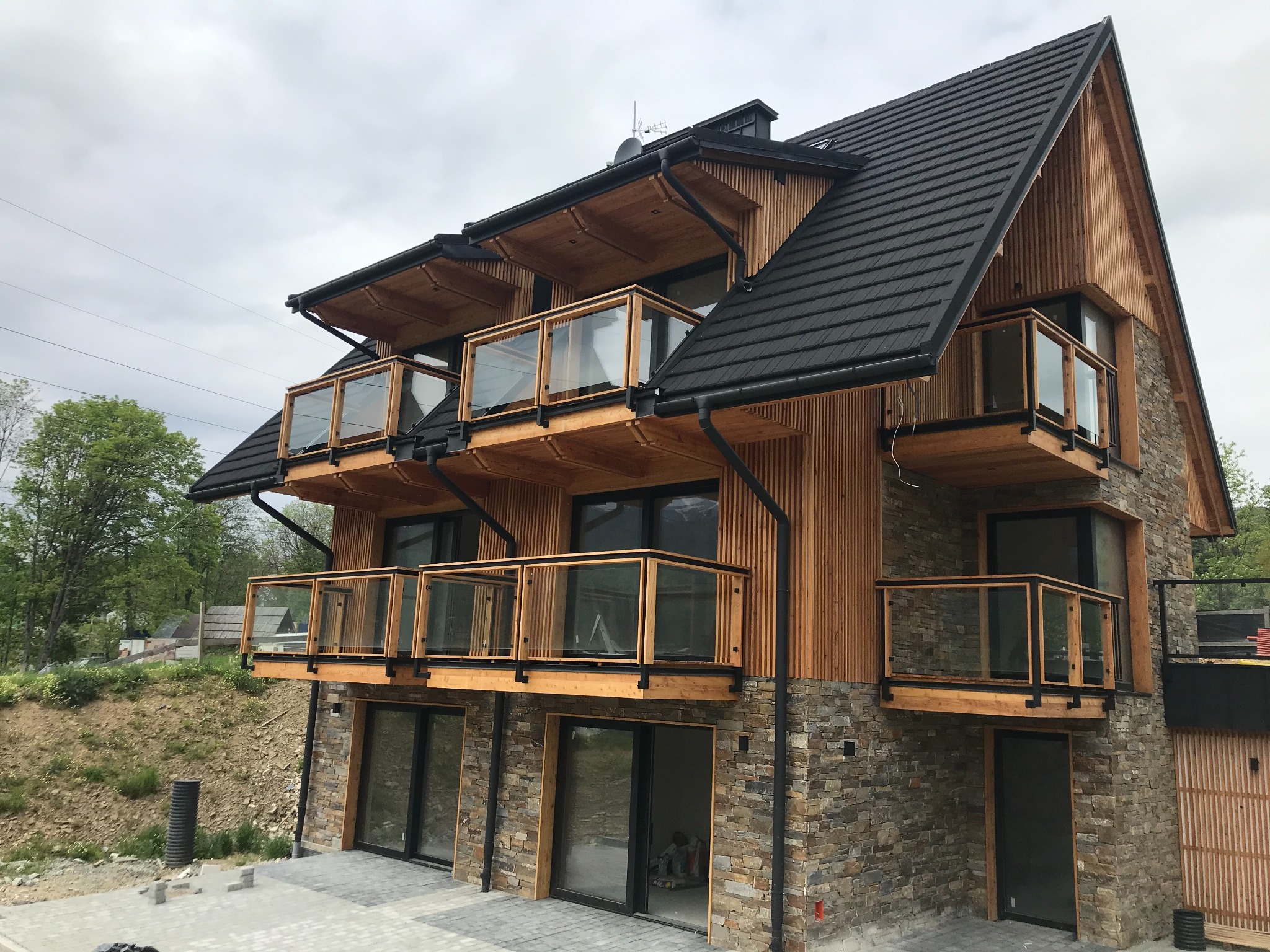 Nowoczesny dom z drewnianą elewacją i kamienną podmurówką, balkony z szklanymi balustradami, czarne rynny i okna, widok z dołu na tle pochmurnego nieba.