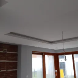 Wykończone wnętrze z podwieszanym sufitem z dekoracyjnymi wgłębieniami, szarą ścianą i fragmentem ściany z cegły, widoczne okna z drewnianą ramą i wisząca żarówka bez klosza.