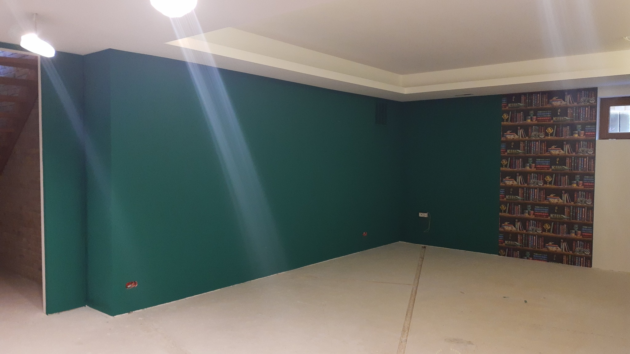 Pokój po malowaniu: ściany w kolorze butelkowej zieleni, jedna ściana z tapetą imitującą regał z książkami, jasna podłoga w trakcie przygotowania, widoczne gniazdka elektryczne.