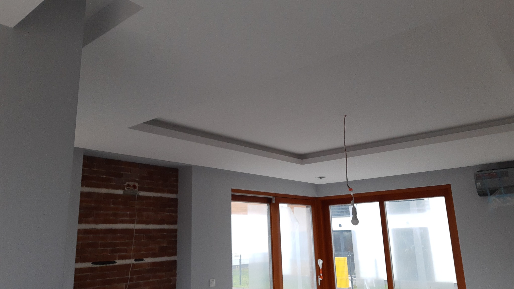 Wykończone wnętrze z podwieszanym sufitem z dekoracyjnymi wgłębieniami, szarą ścianą i fragmentem ściany z cegły, widoczne okna z drewnianą ramą i wisząca żarówka bez klosza.