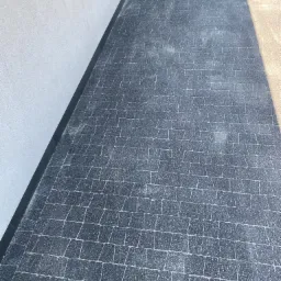 Ułożona nawierzchnia z ciemnoszarych kostek brukowych na tarasie, przylegająca do jasnej ściany budynku z widocznym cieniem.