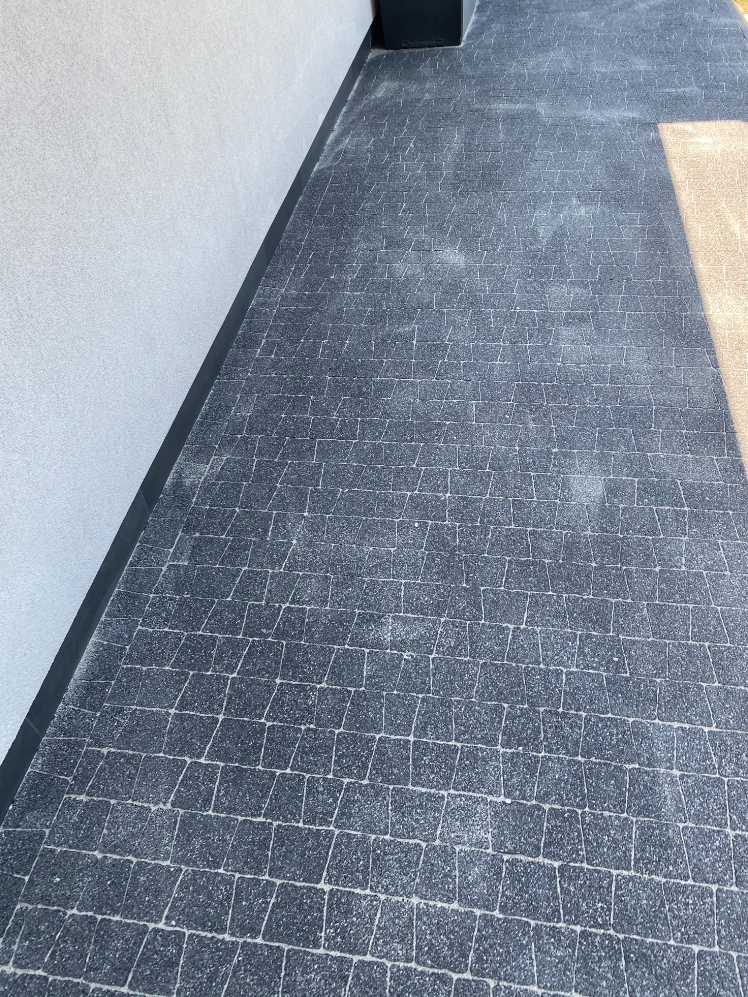 Ułożona nawierzchnia z ciemnoszarych kostek brukowych na tarasie, przylegająca do jasnej ściany budynku z widocznym cieniem.