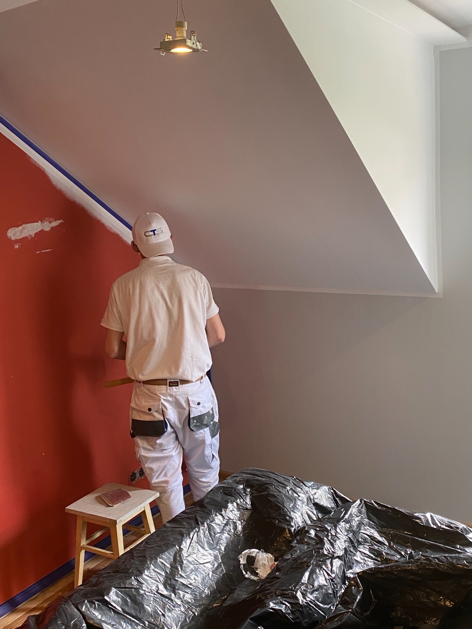 Mężczyzna w białych spodniach i koszulce, z czapką na głowie, maluje czerwoną ścianę na biało, stojąc na tle zabezpieczonych folią mebli.