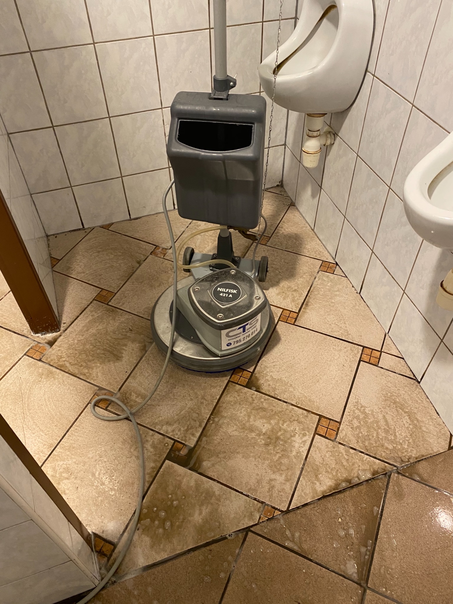 Profesjonalna maszyna czyszcząca Nilfisk 431 A podczas mycia podłogi z płytek w publicznej toalecie z pisuarami i umywalką.