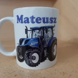 Biały ceramiczny kubek z nadrukiem niebieskiego ciągnika New Holland i imieniem 'Mateusz' w kolorze niebieskim, ustawiony na drewnianej powierzchni.
