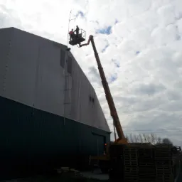 Dwóch pracowników w koszu podnośnika koszowego, na tle pochmurnego nieba, przy ścianie hali magazynowej z blachy trapezowej, obok sterty palet.