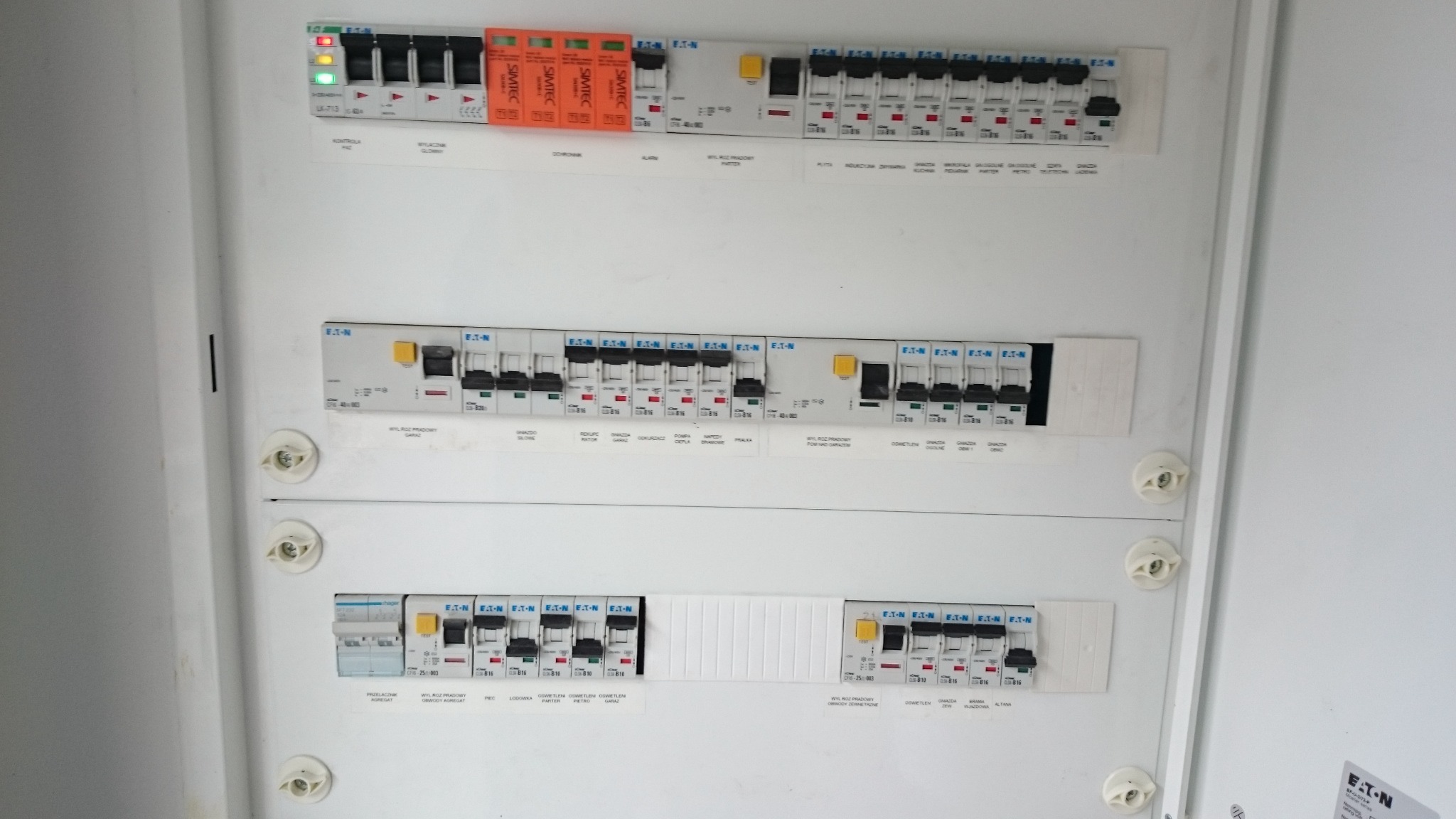Rozdzielnica elektryczna z widocznymi bezpiecznikami i wyłącznikami różnicowoprądowymi marki Eaton oraz Simtec, opisane obwody, zamontowana w białej skrzynce.