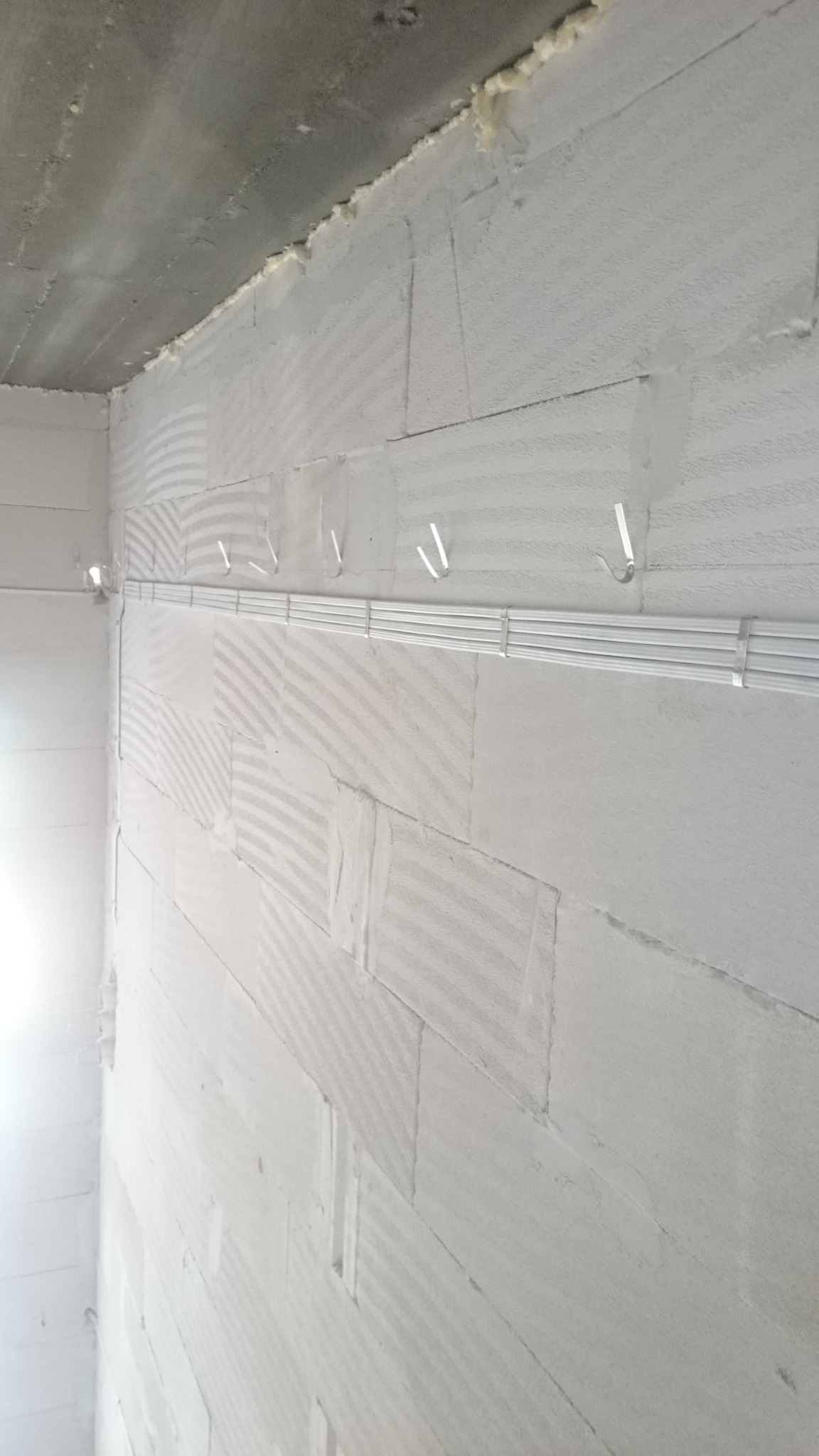 Szara ściana z betonu komórkowego, na której zamontowano koryto kablowe z białymi przewodami elektrycznymi, przymocowane tymczasowymi uchwytami.