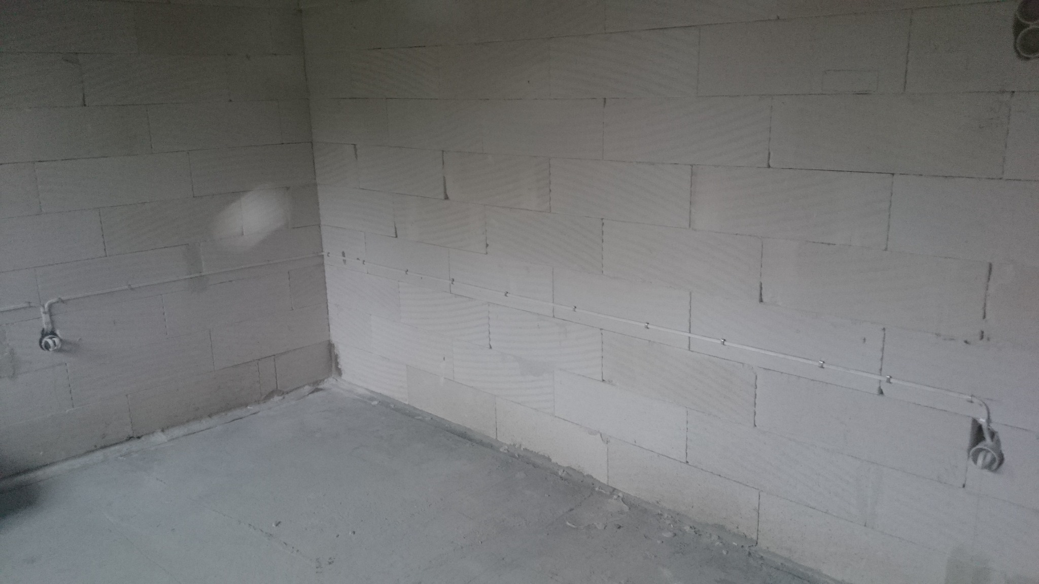 Surowy stan wnętrza z widoczną instalacją elektryczną: kable poprowadzone po ścianie z betonu komórkowego, zamontowane puszki elektryczne w rogach pomieszczenia, betonowa wylewka podłogowa.