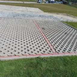 Parking z ażurowych płyt betonowych, oddzielony czerwoną kostką, z widocznymi drobnymi kamieniami w przestrzeniach między płytami, trawa na obrzeżach.