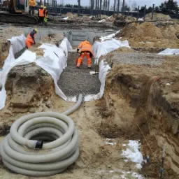 Wykopy pod fundamenty zabezpieczone geowłókniną i wypełnione kruszywem, pracownicy w pomarańczowych kombinezonach, rura drenarska zwinięta w krąg, w tle słupy wysokiego napięcia i koparka.