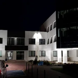 Nowoczesny budynek biurowy z białymi panelami i czarnymi oknami, oświetlony nocą, z widocznym samochodem na pierwszym planie i dekoracyjnymi elementami roślinnymi na elewacji.