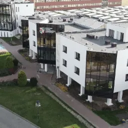 Nowoczesny, biały budynek biurowy z czarnymi, szklanymi fasadami i płaskim dachem, widok z góry na otoczenie z zielenią i parkingiem.