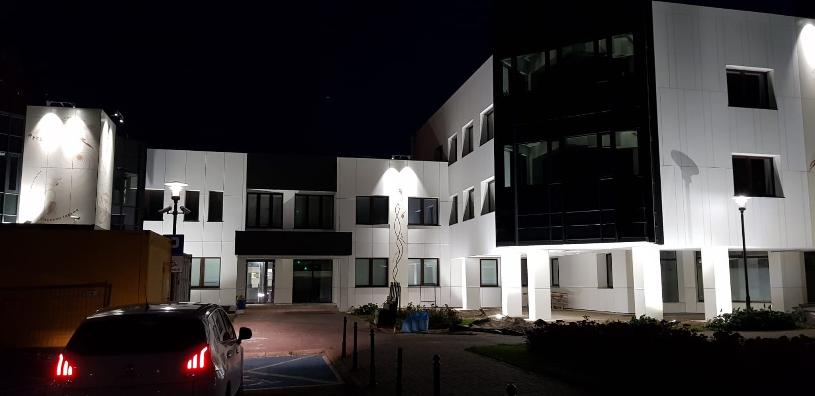 Nowoczesny budynek biurowy z białymi panelami i czarnymi oknami, oświetlony nocą, z widocznym samochodem na pierwszym planie i dekoracyjnymi elementami roślinnymi na elewacji.