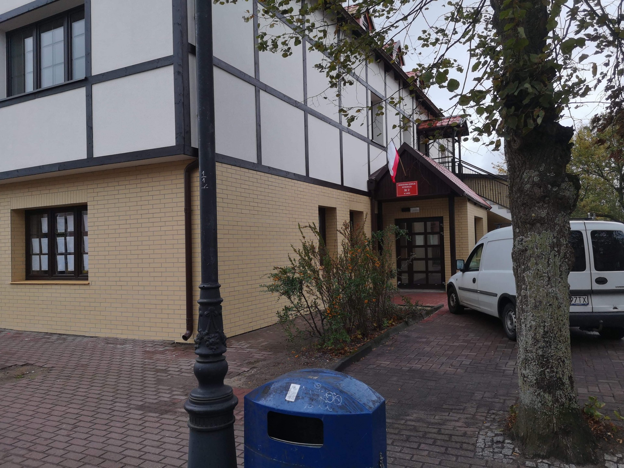 Budynek z murem pruskim, żółtą cegłą i polską flagą nad wejściem, widoczny z zewnątrz, z koszem na śmieci i zaparkowanym białym vanem.