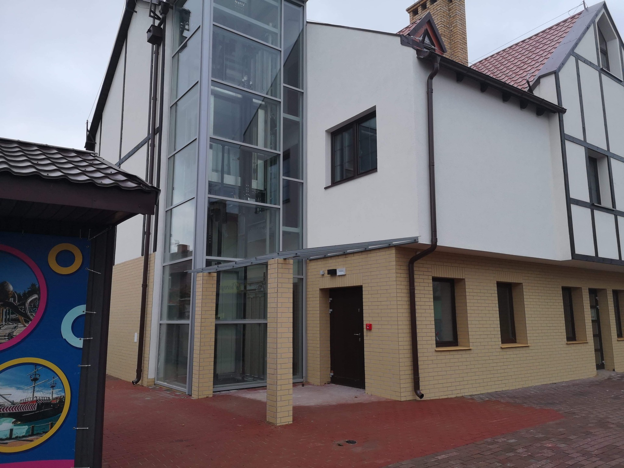 Budynek z przeszkloną windą zewnętrzną, widoczne fragmenty elewacji w stylu szachulcowym, ceglana podmurówka i czerwona kostka brukowa.