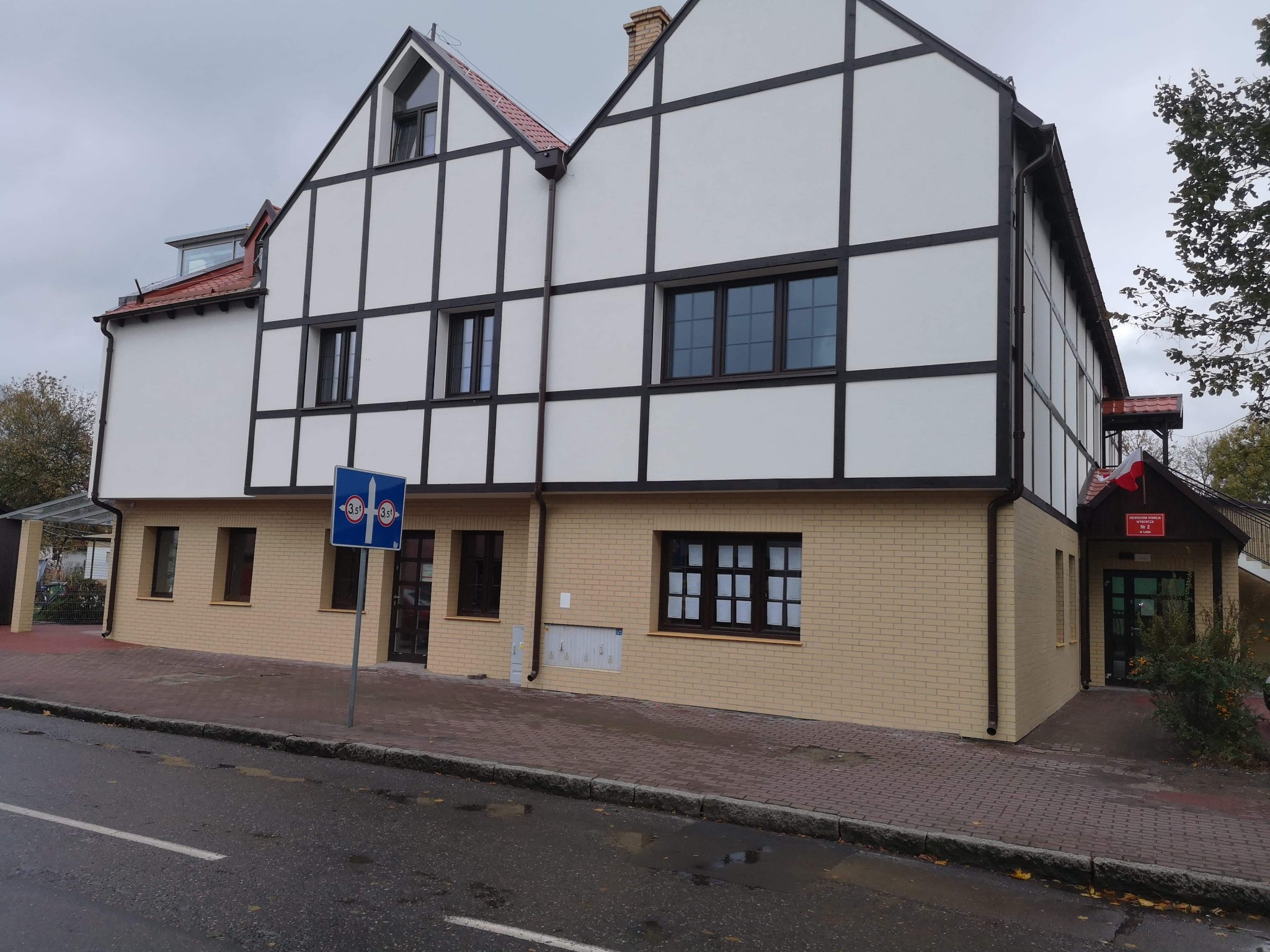 Budynek dwukondygnacyjny z białymi ścianami i ciemnymi belkami w stylu szachulcowym, z żółtą ceglaną podstawą i oknami z ciemnymi ramami, widoczny znak drogowy.