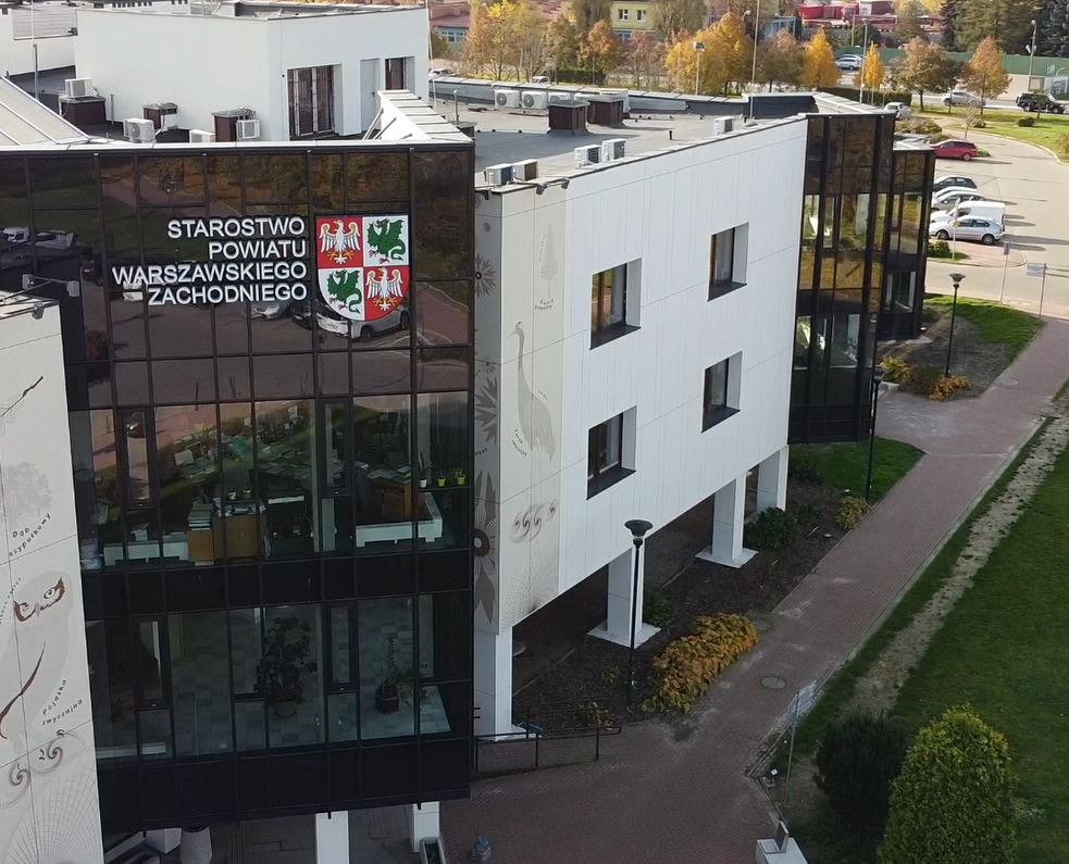 Nowoczesny budynek Starostwa Powiatu Warszawskiego Zachodniego z herbem na szklanej fasadzie i rysunkami ptaków na białej elewacji.