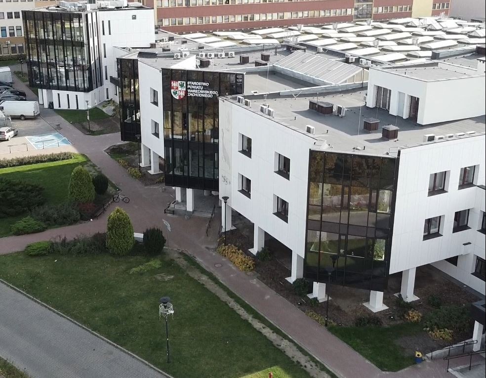 Nowoczesny, biały budynek biurowy z czarnymi, szklanymi fasadami i płaskim dachem, widok z góry na otoczenie z zielenią i parkingiem.