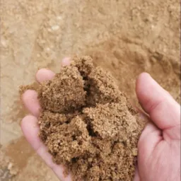 Dłoń trzymająca wilgotny, brązowy piasek frakcji średniej na tle hałdy tego samego piasku.