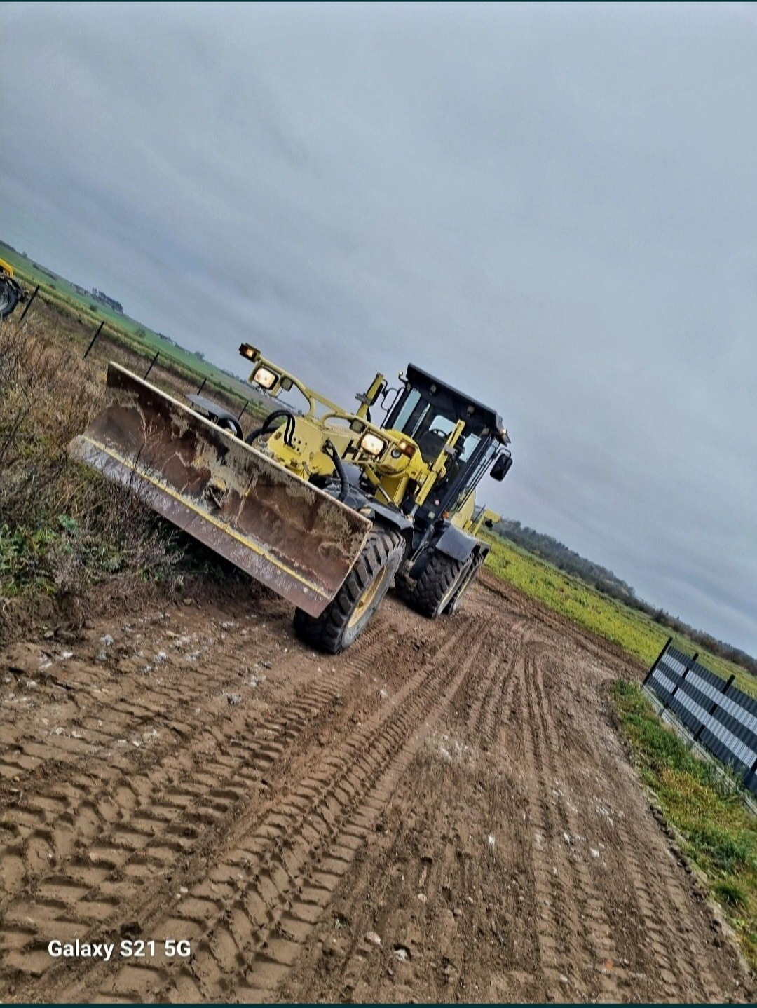 Żółty równiarka na błotnistym terenie z wyraźnymi śladami opon, widoczne pole w tle pod pochmurnym niebem.