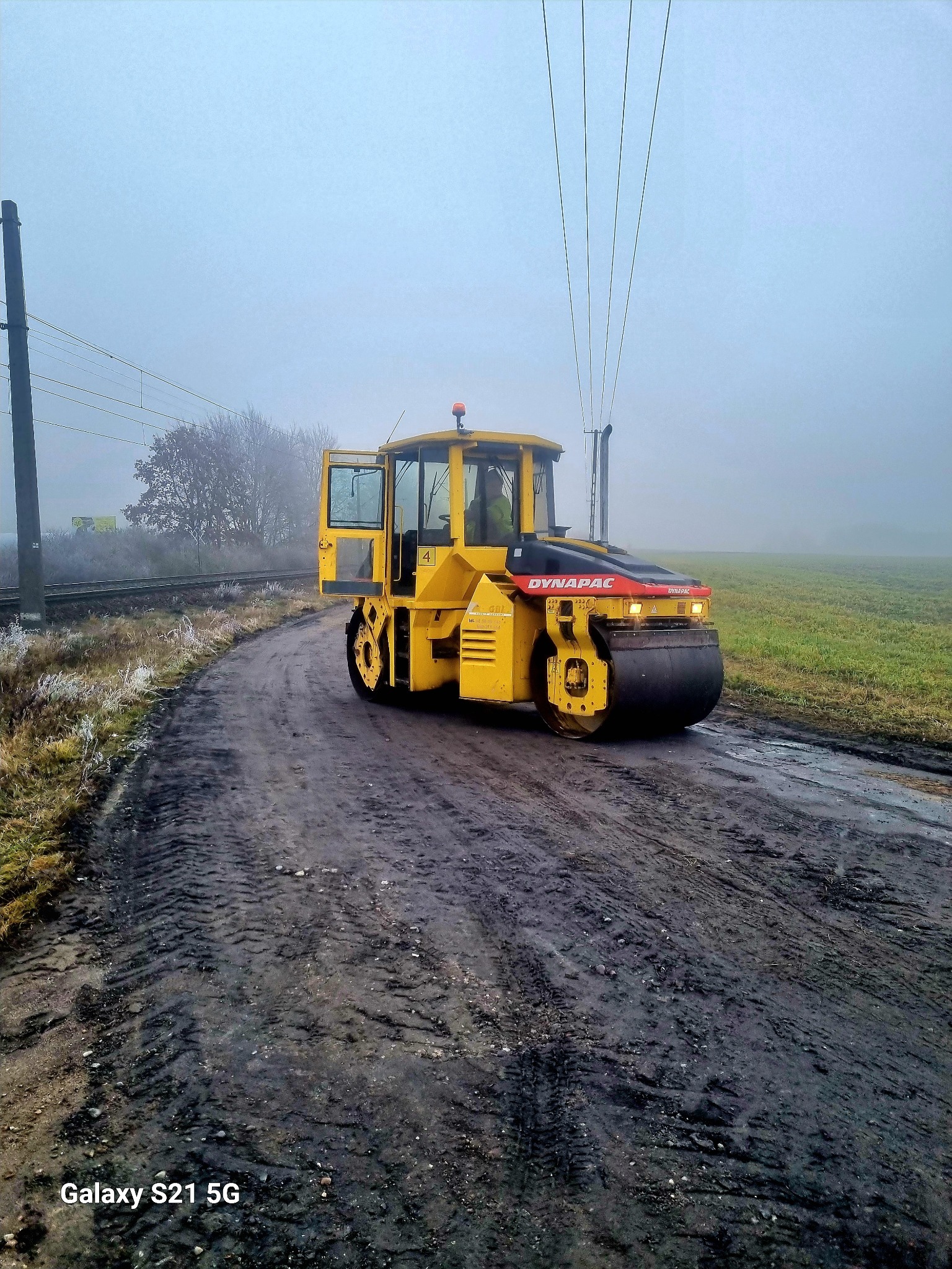 Żółty walec Dynapac ugniata błotnistą drogę gruntową w mglisty dzień, widoczne tory kolejowe w tle.