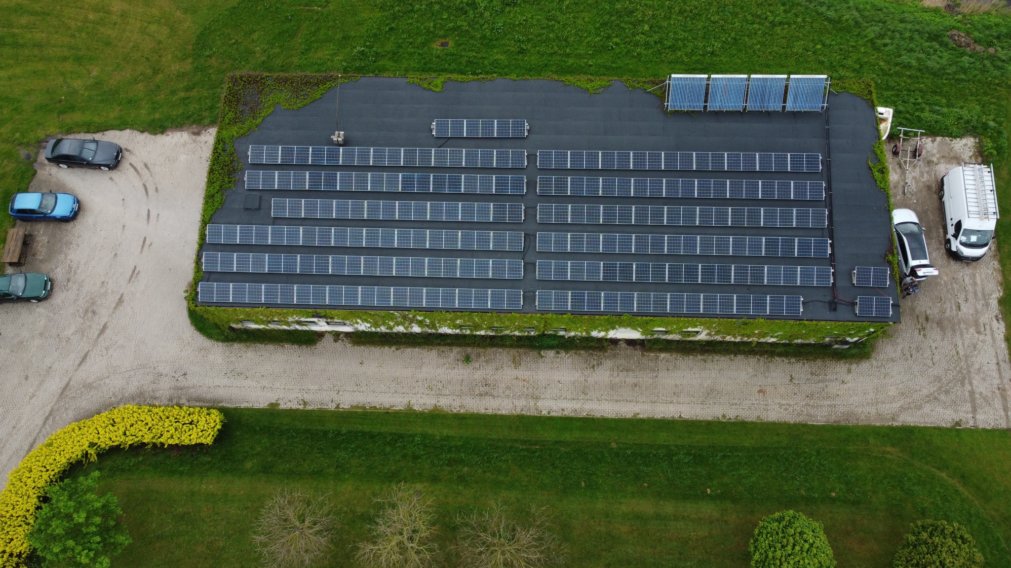 Widok z lotu ptaka na budynek z panelami słonecznymi na dachu, zarośnięty bluszczem, obok zaparkowane samochody i furgonetka z drabiną.