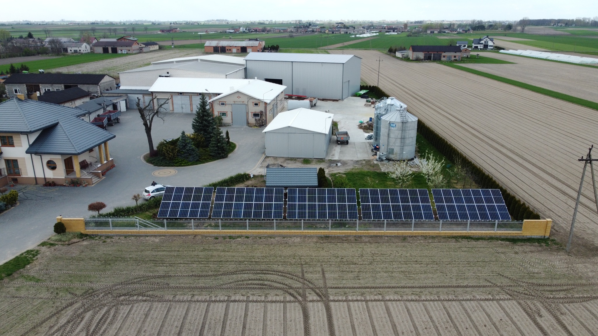 Widok z lotu ptaka na instalację paneli słonecznych w gospodarstwie rolnym, z budynkami gospodarczymi, silosami, domem mieszkalnym i polami uprawnymi w tle.