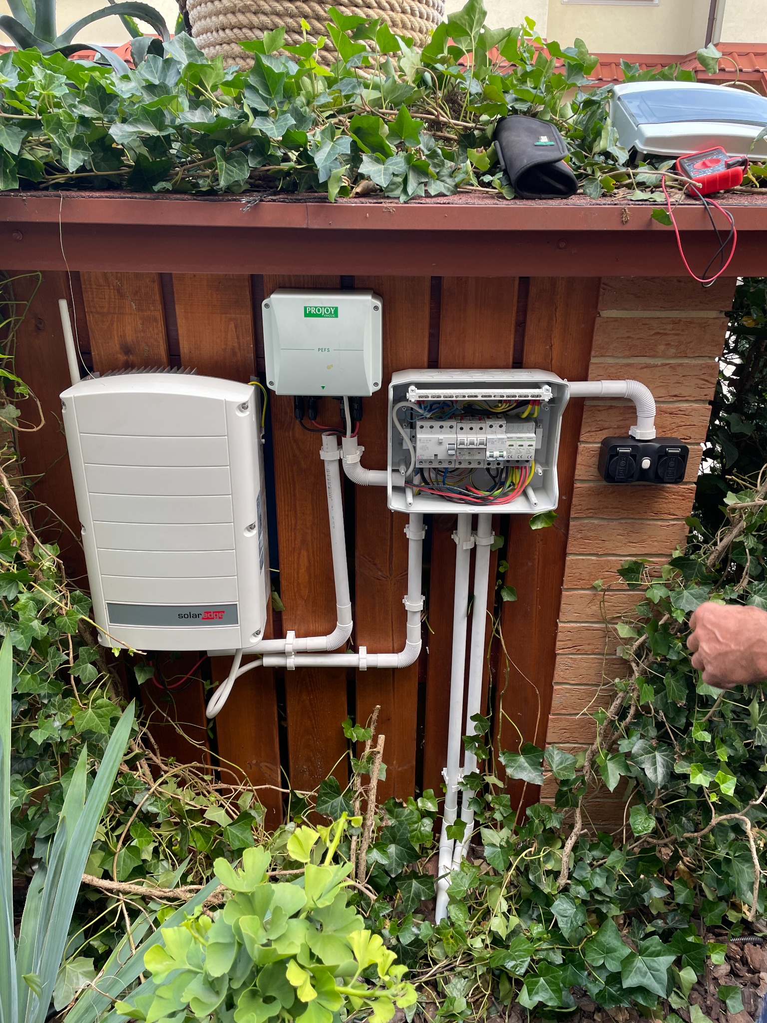 Instalacja elektryczna systemu solarnego z falownikiem SolarEdge, skrzynką bezpieczników i dodatkowymi elementami, przymocowana do drewnianego ogrodzenia, otoczona zielenią pnącą.