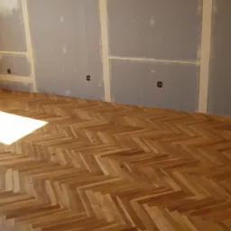 Parkiet ułożony w jodełkę, świeżo po położeniu, z widocznymi śladami szpachlowania na ścianach i wystającymi gniazdkami elektrycznymi.