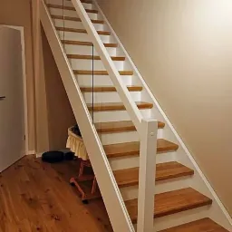 Nowoczesne schody drewniane z białymi balustradami i szklanymi panelami, prowadzące na strych; widoczna podłoga z desek i fragment drzwi.