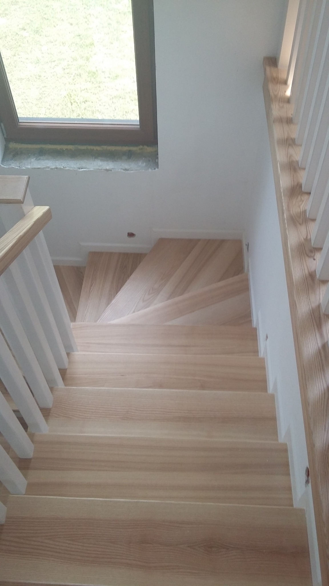 Jasne, drewniane schody z białymi balustradami, widok z góry, z oknem dachowym w tle, ukazujące naturalne usłojenie drewna.