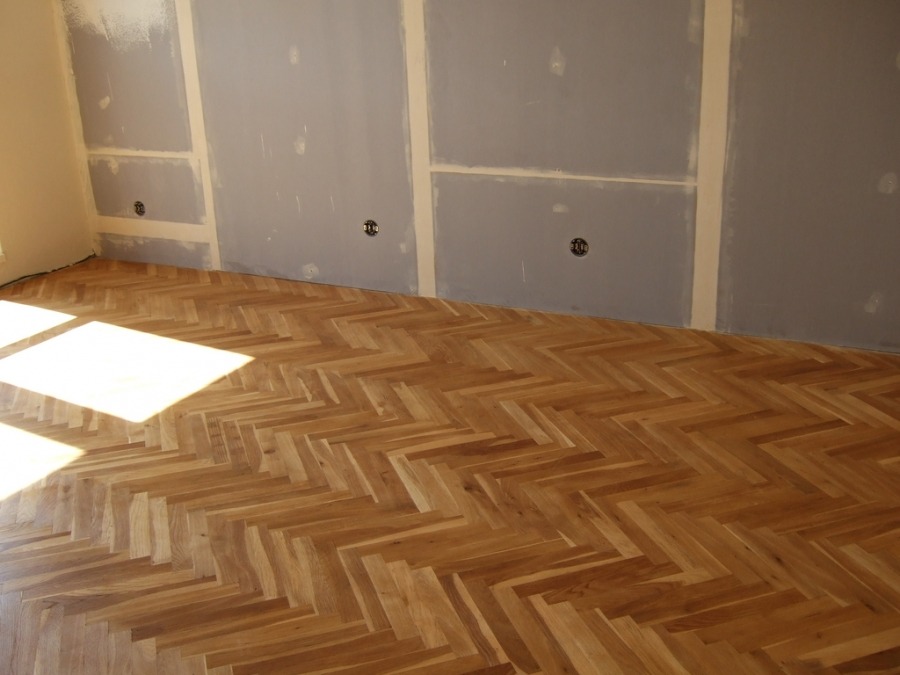 Parkiet ułożony w jodełkę, świeżo po położeniu, z widocznymi śladami szpachlowania na ścianach i wystającymi gniazdkami elektrycznymi.