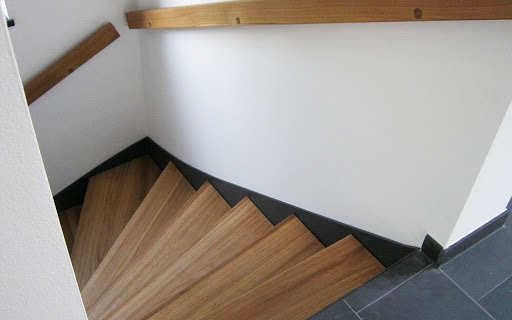 Schody drewniane z jasnego drewna, widok z góry, z ciemnymi listwami przypodłogowymi i fragmentem drewnianej poręczy.