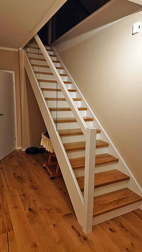 Nowoczesne schody drewniane z białymi balustradami i szklanymi panelami, prowadzące na strych; widoczna podłoga z desek i fragment drzwi.