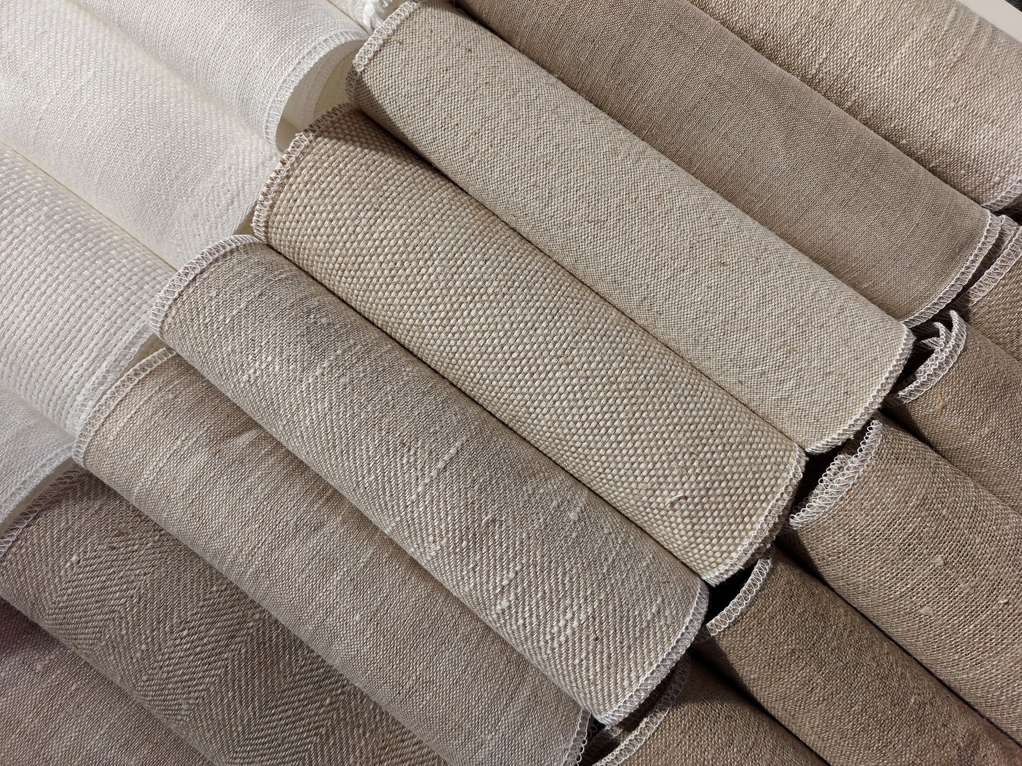 Stos zwiniętych rolek tkanin o różnej fakturze i odcieniach beżu, z widocznymi brzegami zabezpieczonymi overlockiem.