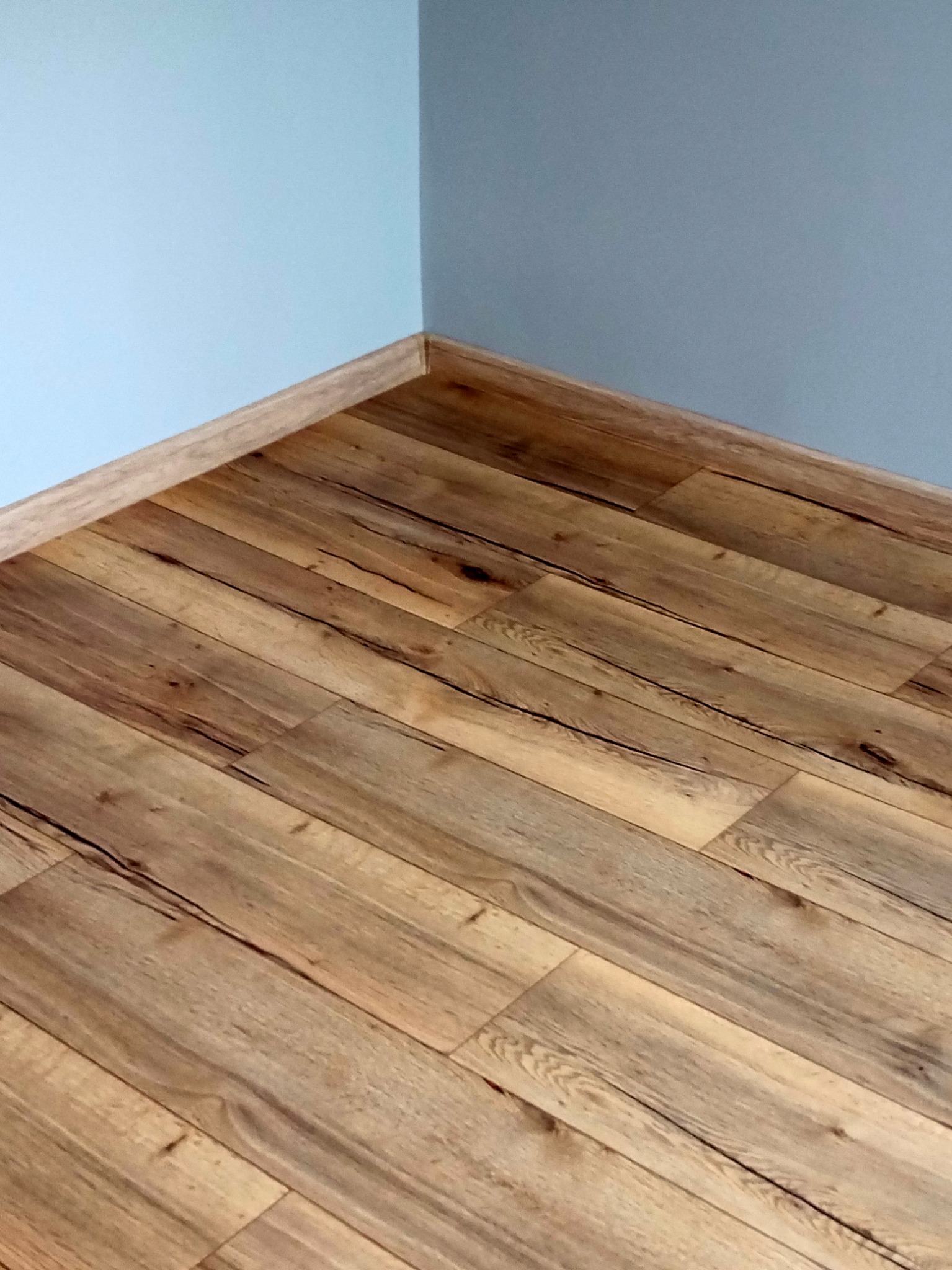 Fragment pokoju z drewnianą podłogą ułożoną w jodełkę, jasnoniebieskie ściany i listwa przypodłogowa w kolorze drewna.