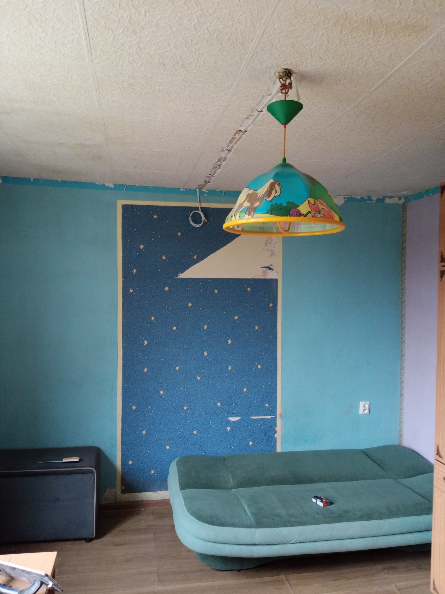 Pokój przed remontem: niedokończone ściany z resztkami tapet w gwiazdki, niebieskie ściany z ubytkami farby, sufit z widocznymi łączeniami płyt i wisząca lampa z motywem zwierząt, sofa w kolorze...