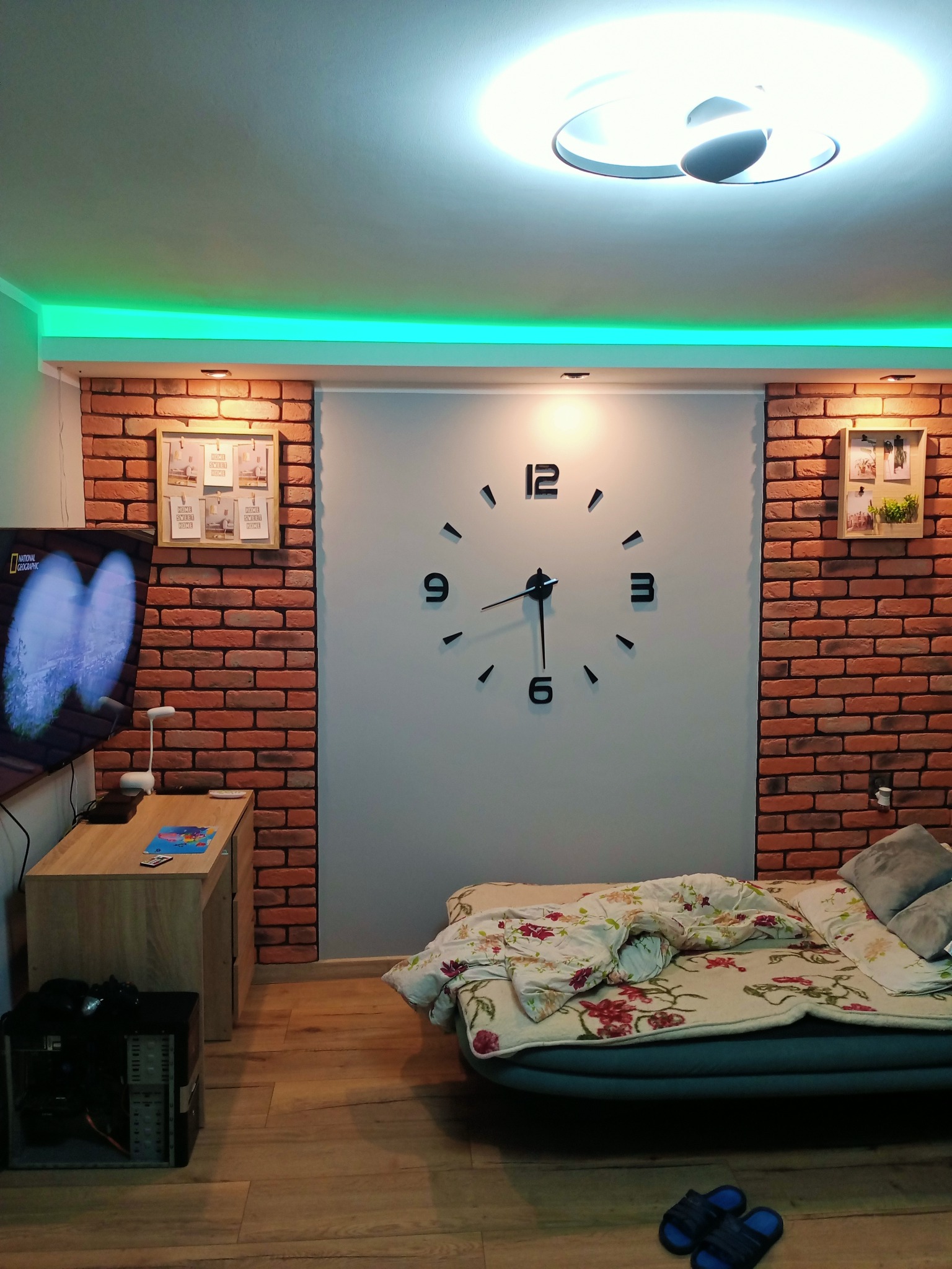 Pokój z łóżkiem na podłodze, ceglaną ścianą, dużym zegarem ściennym i podświetleniem LED na suficie.