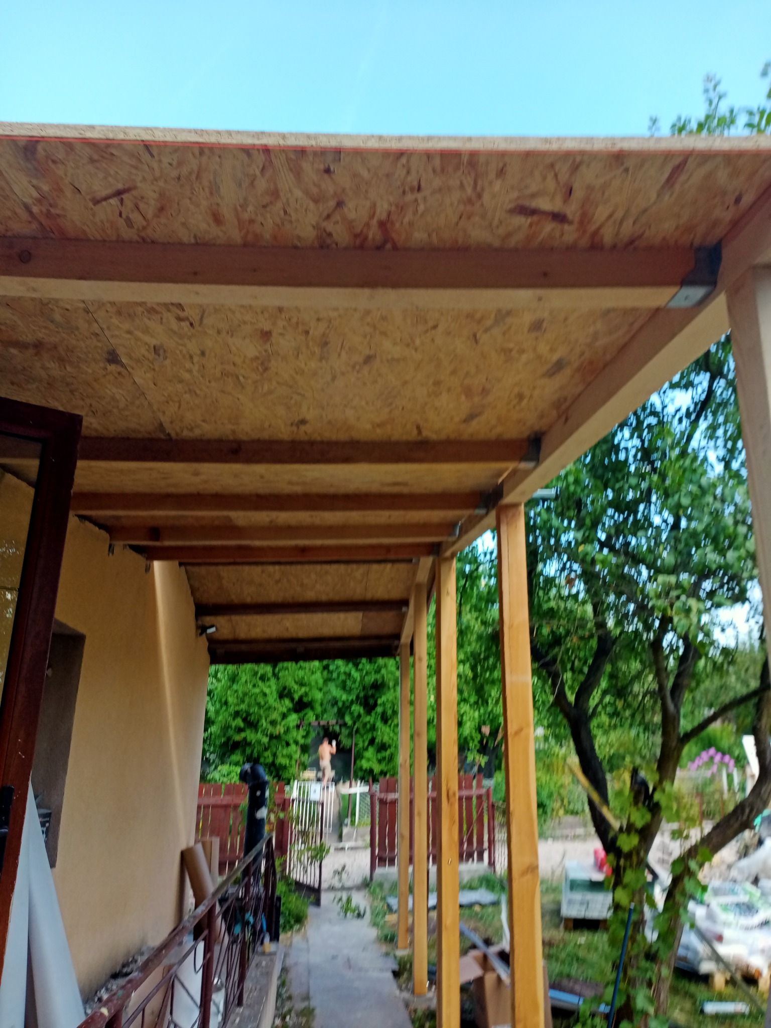 Zadaszenie tarasu w trakcie budowy: drewniane belki nośne połączone metalowymi wspornikami, pokryte płytą OSB, widok z dołu na konstrukcję i otoczenie ogrodu.