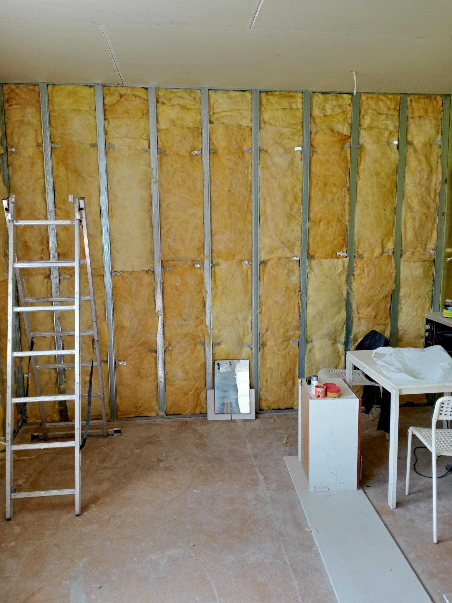 Ściana z metalowym stelażem wypełnionym żółtą wełną mineralną, widoczna drabina aluminiowa, fragment białego stolika i krzesła oraz małe lusterko na podłodze w remontowanym pomieszczeniu.
