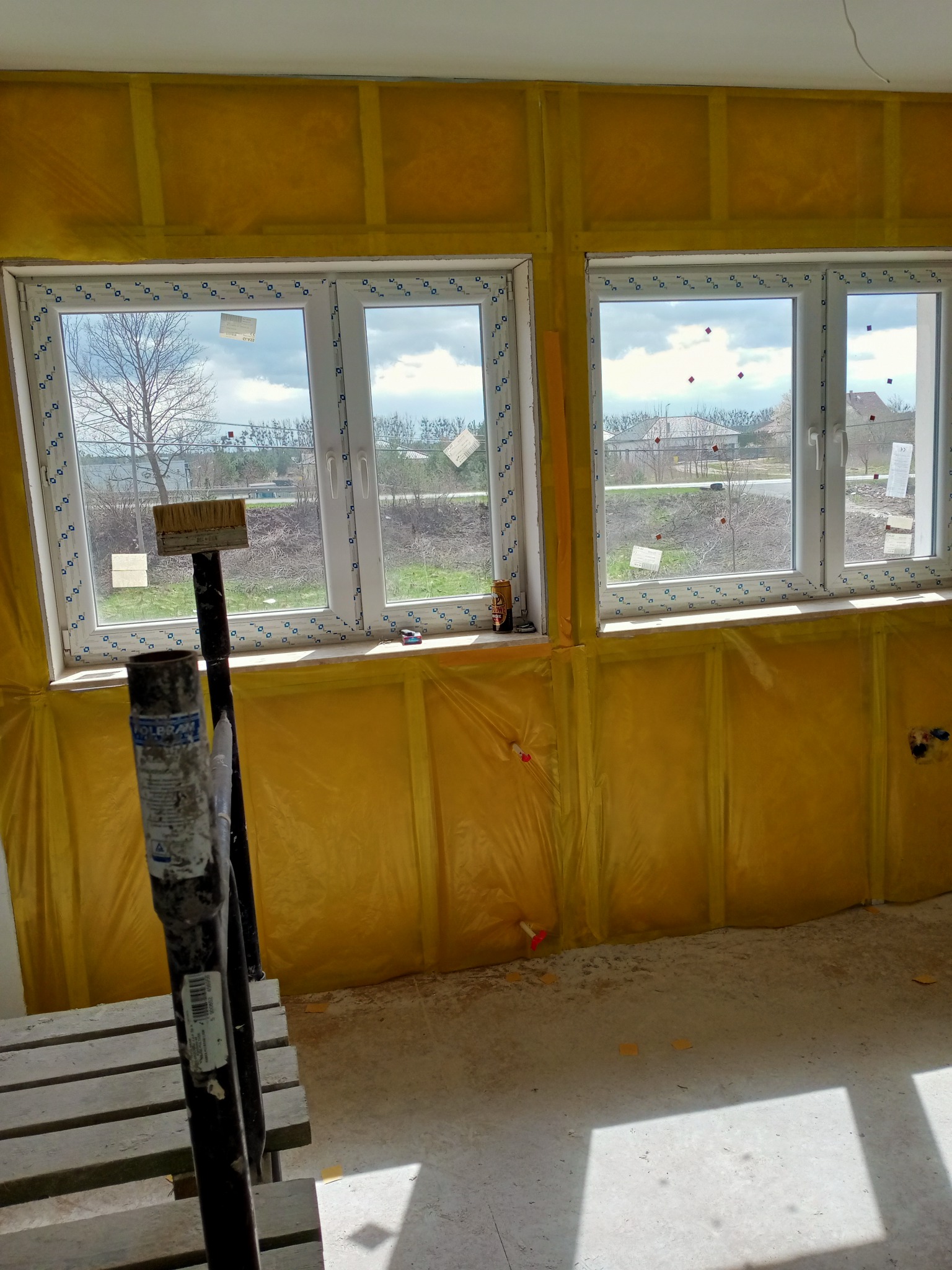 Dwa białe okna PCV w surowym wnętrzu, ściany pokryte żółtą folią ochronną, widoczny fragment drabiny malarskiej i pędzla, przygotowanie do prac wykończeniowych.