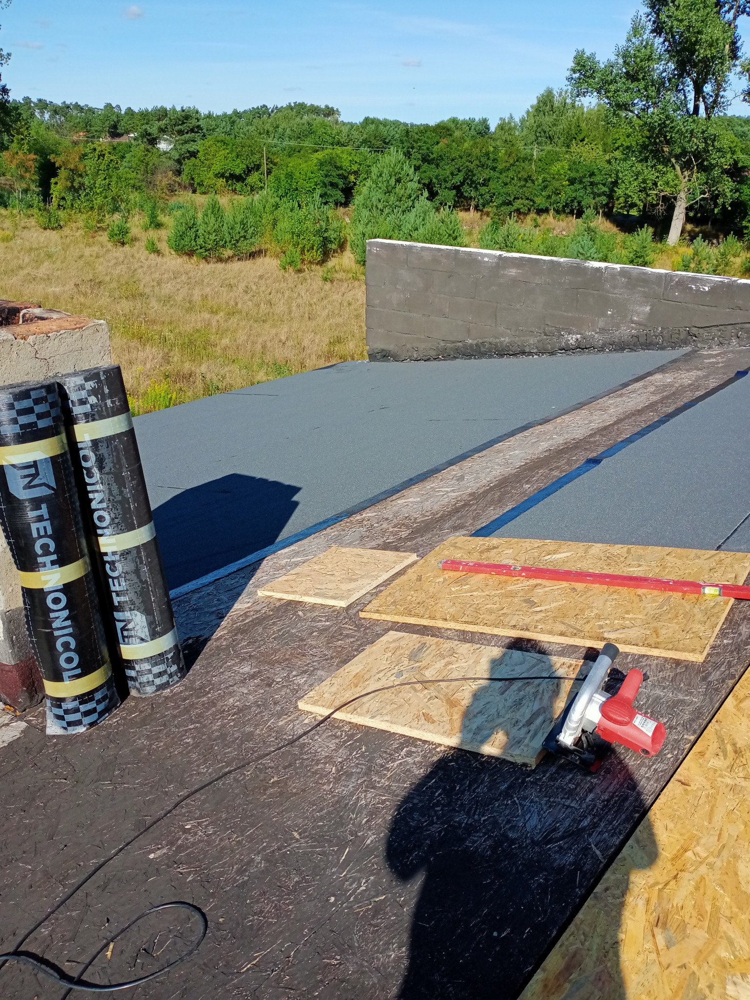 Prace na dachu: rolki papy termozgrzewalnej Technonicol, płyty OSB, poziomica i piła elektryczna na tle zieleni.